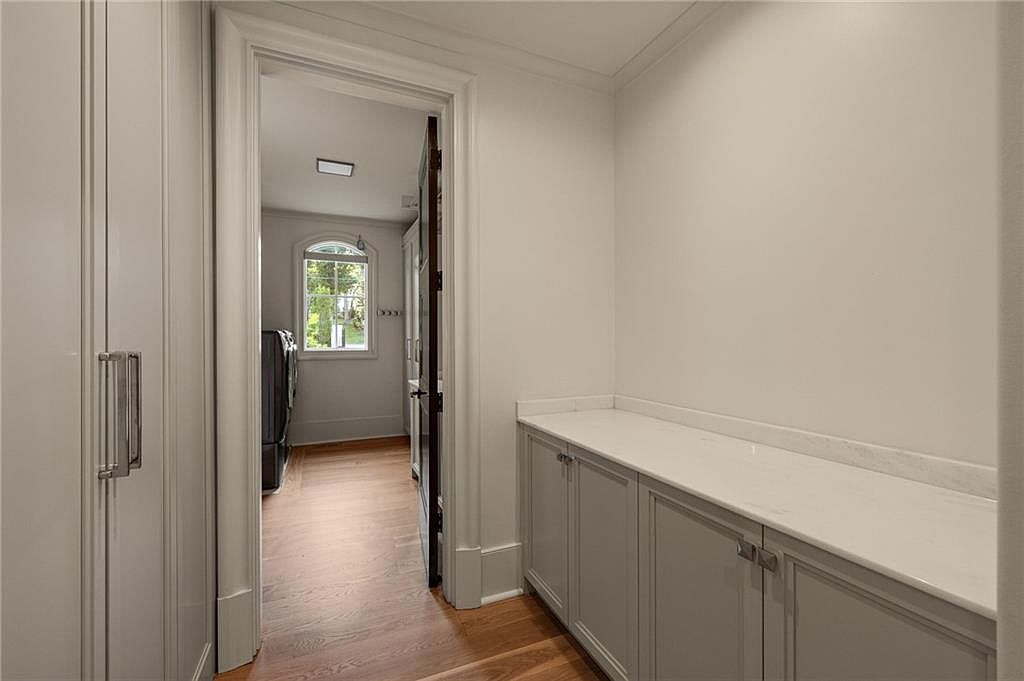 This view captures a transition space featuring light-colored cabinetry with a smooth countertop, leading through a doorway into a laundry area. The perspective looks from a hallway with hardwood flooring toward a bright room containing a washer and dryer set near a window. The neutral color palette and clean lines create a cohesive and functional aesthetic throughout the home.