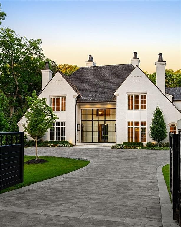 This elegant, white-painted brick residence showcases a modern take on traditional architecture with a steep, dark-shingled roof and symmetrical gables. A prominent central entryway features expansive, grid-patterned glass panels that extend from the ground to the roofline, creating a striking focal point. The property is approached by a wide, paved driveway flanked by manicured green lawns and mature trees, conveying a sense of luxury and privacy.