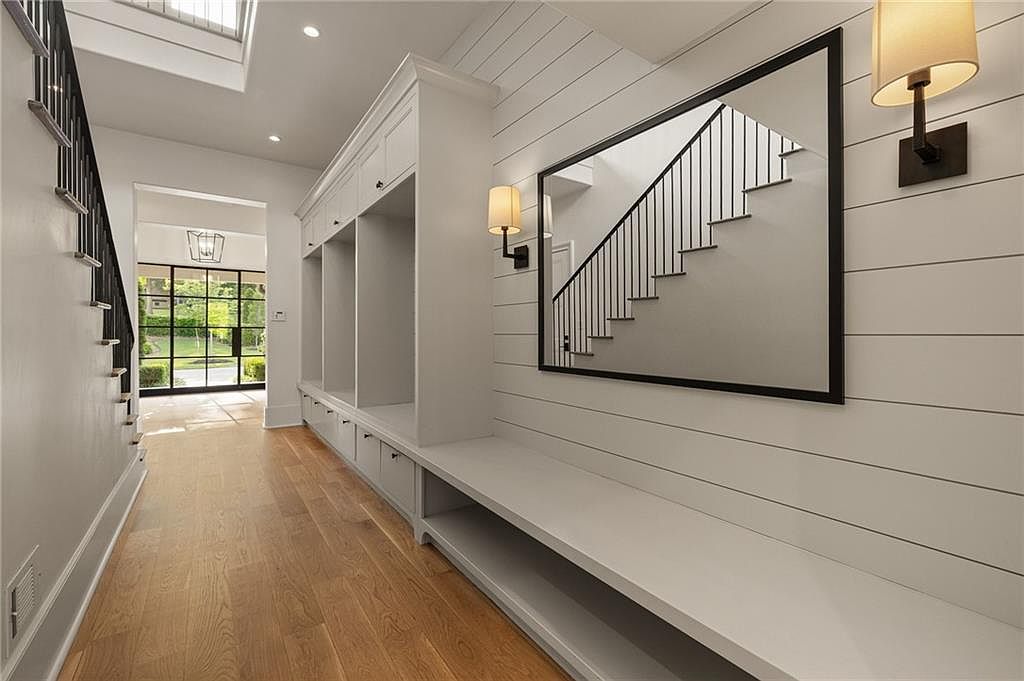 This bright and modern entryway features a long built-in mudroom bench with integrated storage cabinets and cubbies. The walls are adorned with horizontal white shiplap, complemented by a large rectangular mirror that reflects the adjacent staircase. Light-toned wood flooring runs throughout the space, leading toward a glass-paned door that provides a view of the outdoor greenery.