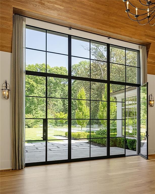 This interior space features a dramatic wall of floor-to-ceiling black-framed windows that provide an expansive view of the lush green backyard and patio. The room is finished with light wood flooring and a matching wood-paneled ceiling, creating a warm and cohesive aesthetic. Elegant wall-mounted lanterns and a partial view of a chandelier add sophisticated lighting details to this bright, airy living area.