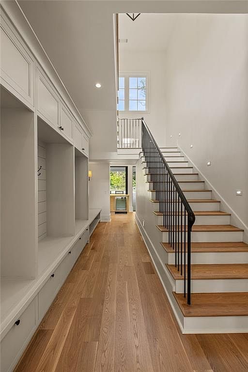 This bright and airy entryway features a long hallway with light wood flooring and a built-in mudroom storage unit with bench seating on the left. A staircase with a black metal railing and light wood treads leads to an upper landing, while a view through to the back of the home reveals a small beverage center. The space is characterized by a clean, modern aesthetic with crisp white walls and recessed lighting.