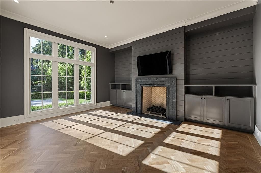 This elegant living room features a sophisticated dark gray shiplap accent wall centered around a stone fireplace with a herringbone-patterned firebox. Large, multi-pane windows flood the space with natural light, highlighting the intricate herringbone-patterned wood flooring. Built-in cabinetry on either side of the fireplace provides ample storage, creating a balanced and polished aesthetic.