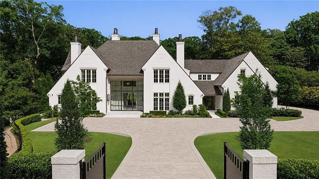 This grand, white-painted brick estate features a symmetrical architectural design with a prominent central entryway framed by large, multi-paned windows. The home is set back from a paved circular driveway, surrounded by lush green lawns and mature trees that provide a sense of privacy and elegance. The perspective is a straight-on, eye-level shot that captures the imposing scale and refined curb appeal of the property.