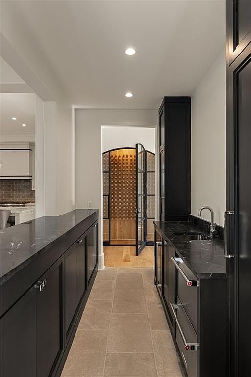 This interior view captures a sophisticated butler's pantry or wet bar area leading toward a glass-enclosed wine cellar. The space features dark cabinetry with stone countertops, a stainless steel sink, and a neutral stone floor, creating a high-end, moody aesthetic. The perspective looks down the narrow corridor, highlighting the transition from the functional bar area to the illuminated wine storage beyond.