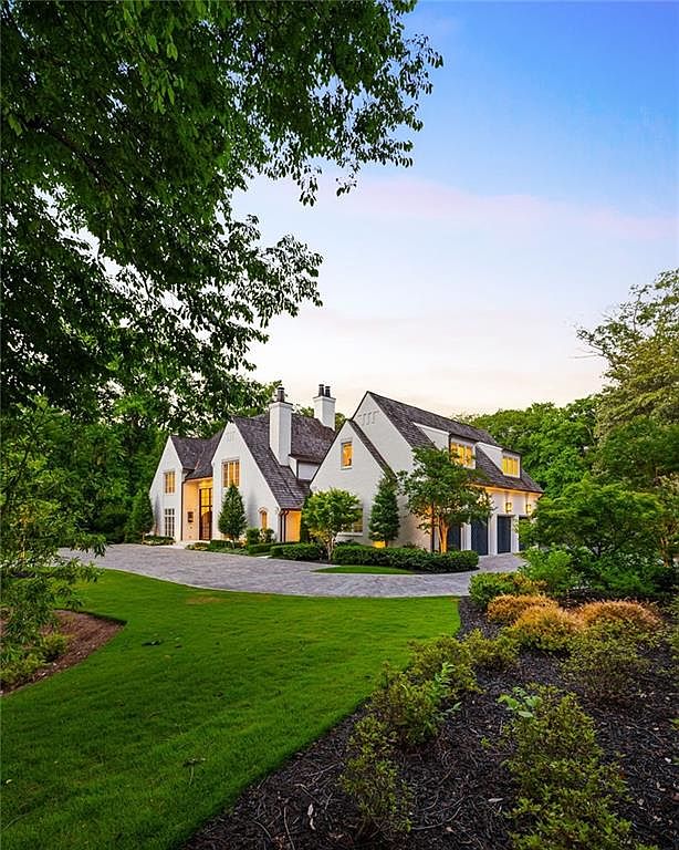 This elegant, multi-gabled estate features a classic white brick exterior with a steep, dark shingled roof, set against a backdrop of lush, mature trees. A wide, paved driveway curves gracefully toward the home, leading to a multi-car garage on the right, while a manicured lawn and landscaped garden beds frame the foreground. The perspective is captured from a low angle, emphasizing the home's grand scale and inviting, serene atmosphere during the golden hour.