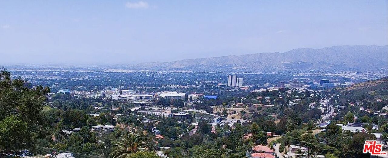 This aerial view showcases a sprawling cityscape nestled among lush greenery and rolling hills. The scene captures a mix of residential areas, commercial buildings, and distant mountain ranges under a clear sky. The perspective provides a sense of the property's location within a vibrant and scenic environment.