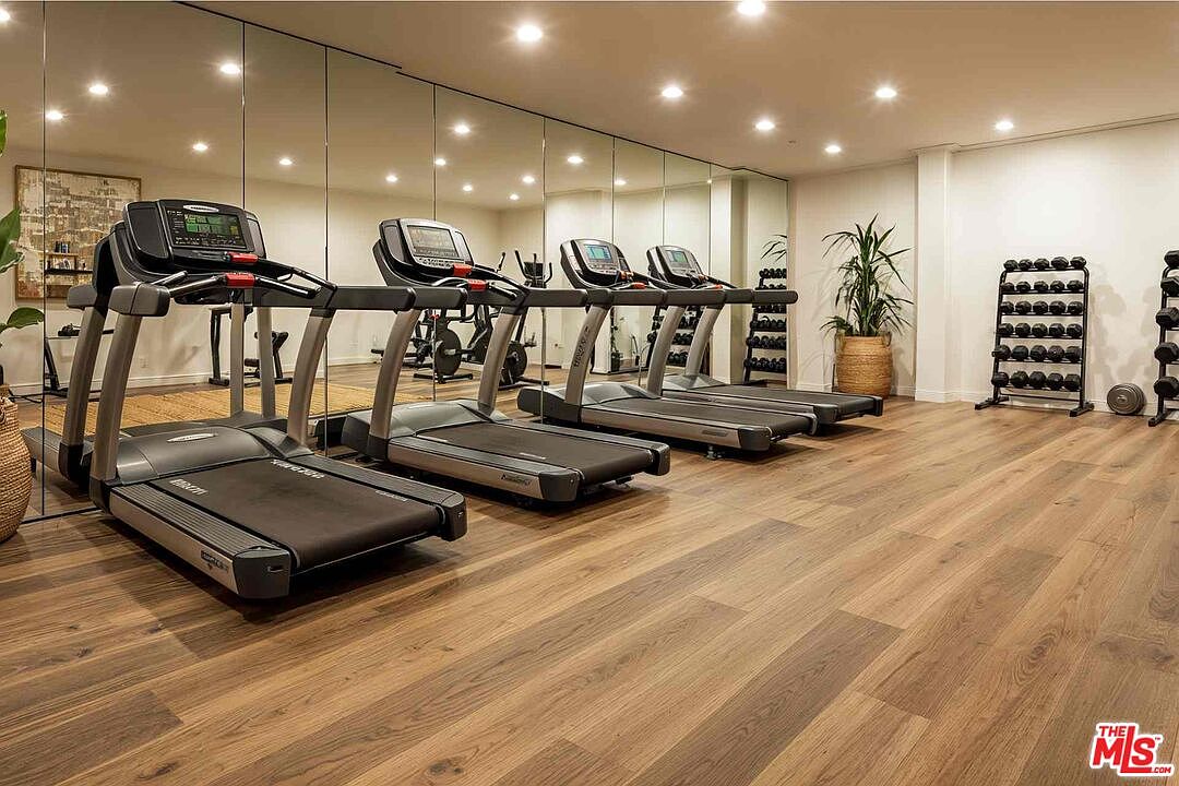 This is a well-equipped home gym featuring multiple treadmills lined up in front of a mirrored wall, creating a sense of spaciousness. A rack of dumbbells is visible on the right, suggesting a comprehensive workout space. The wood flooring adds a touch of warmth to the room.