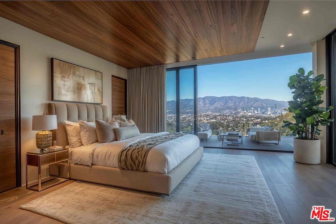 This is a luxurious primary bedroom featuring a king-size bed with a tufted headboard, a modern nightstand with a lamp, and a large abstract painting above the bed. The room boasts floor-to-ceiling windows offering panoramic views of the city and mountains, with access to an outdoor balcony area. The wood-paneled ceiling and neutral color palette create a serene and sophisticated atmosphere.