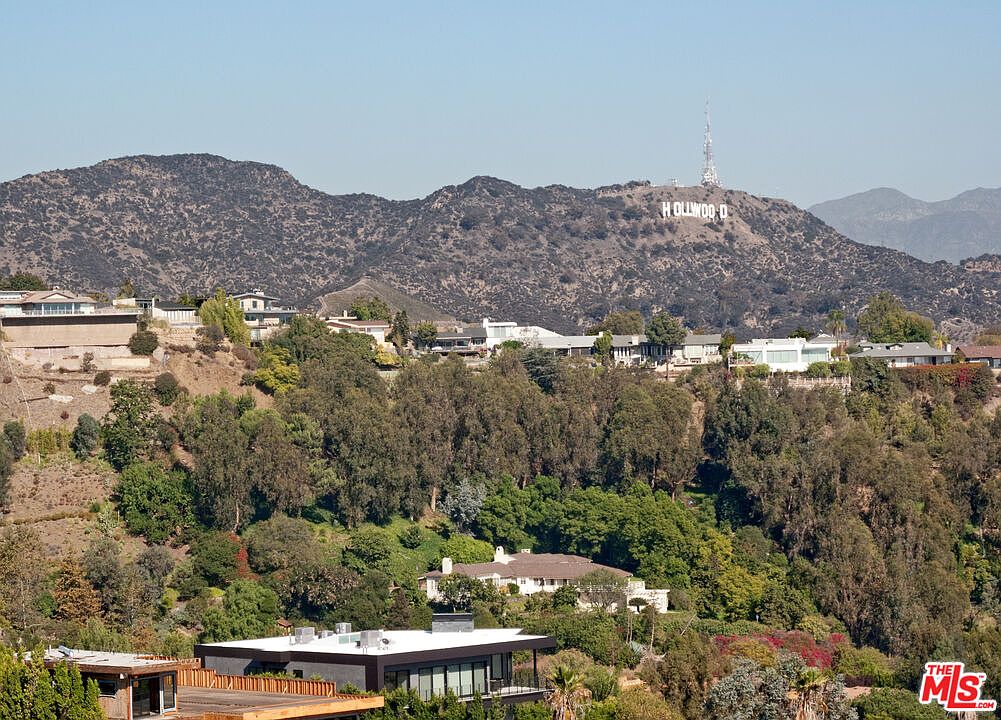 This aerial view showcases a hillside neighborhood with modern homes nestled among lush greenery. The iconic Hollywood sign is visible in the background, adding to the property's appeal. The landscape features mature trees and well-maintained gardens, creating a serene and private setting.