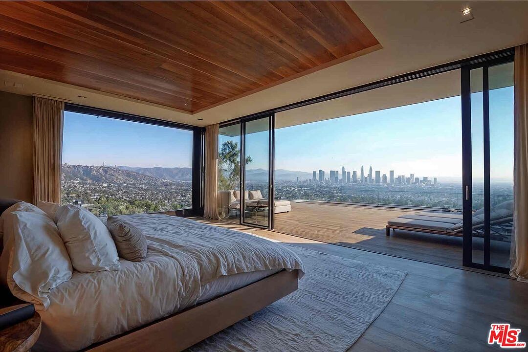 This is a luxurious primary bedroom featuring a large bed with white linens, a wood-paneled ceiling, and expansive sliding glass doors that open to a balcony with city views. The room is spacious and well-lit, creating a serene and upscale atmosphere. The view of the city skyline is a major selling point.