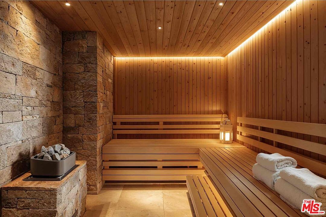 This is an interior shot of a sauna, featuring wooden benches and paneling that create a warm and inviting atmosphere. A stone wall adds a rustic touch, while soft lighting enhances the relaxing ambiance. The sauna includes a heater with stones, suggesting a traditional sauna experience.