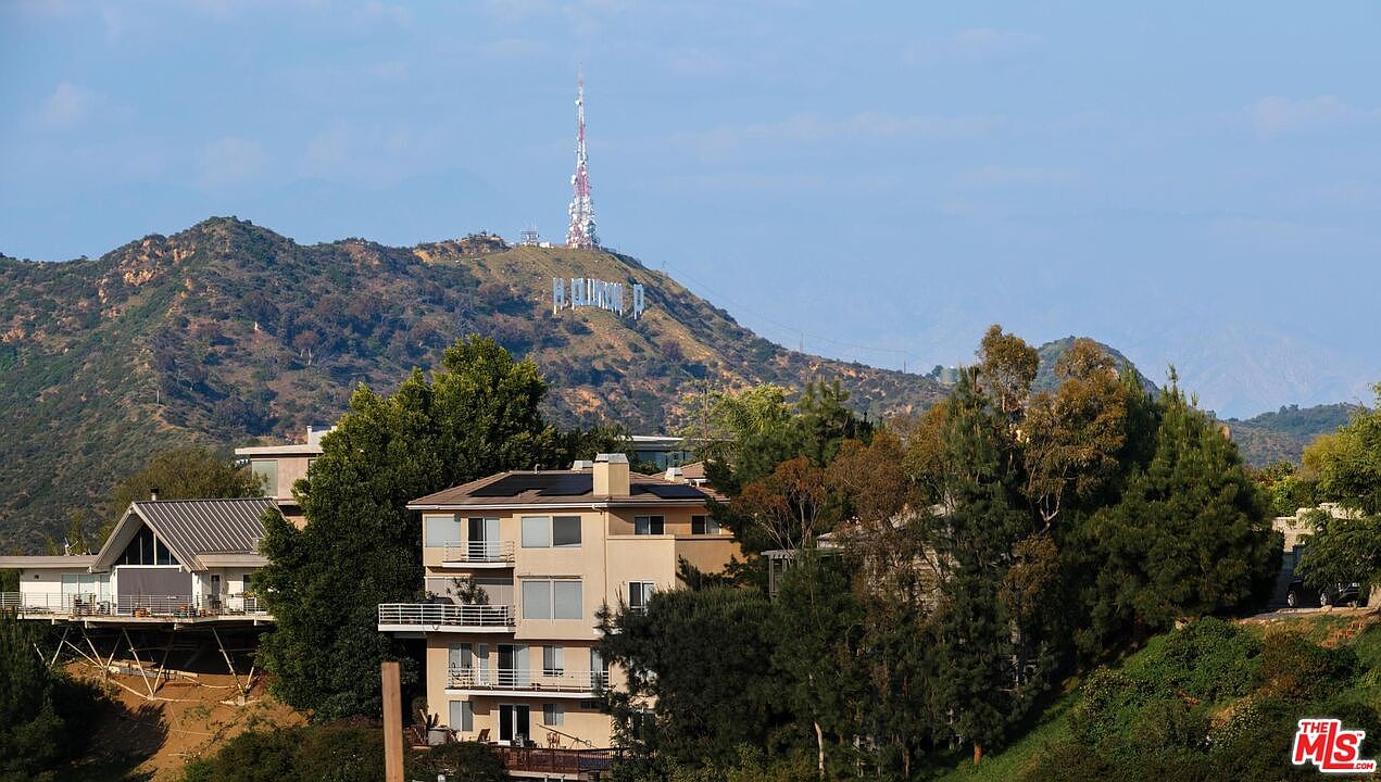 This image showcases the front exterior of a multi-story residential building, possibly a condominium or apartment complex, nestled among lush greenery. The building features balconies and a neutral color palette, blending with the natural surroundings. In the background, the iconic Hollywood sign is visible on a distant hillside, adding a touch of prestige and location appeal.