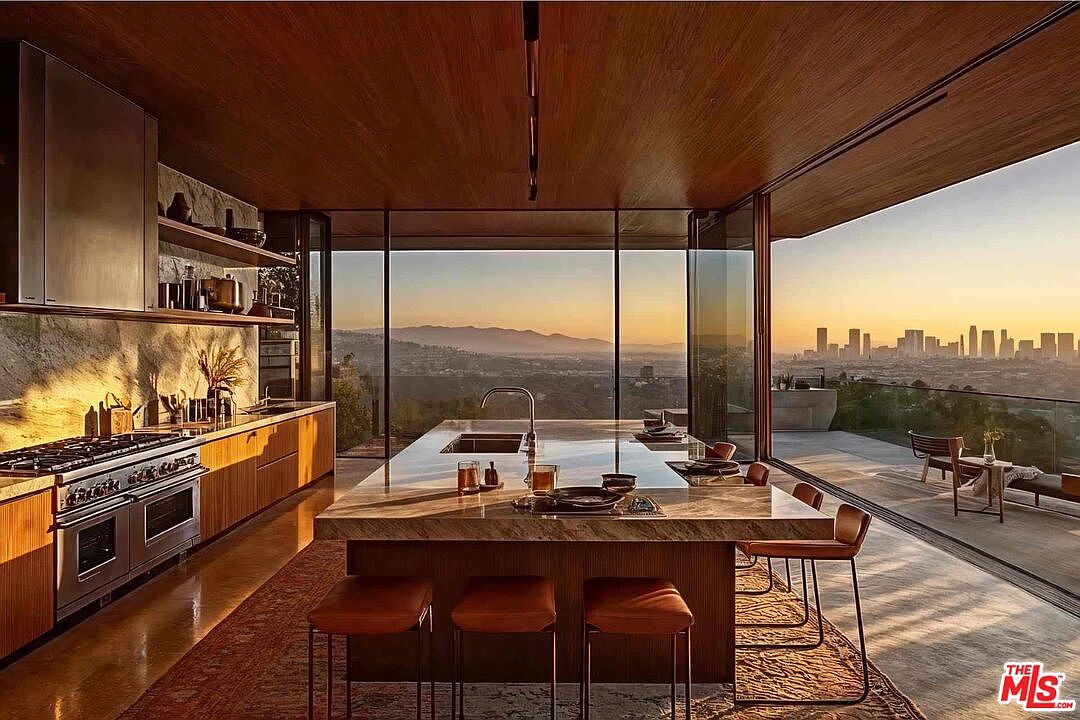 This is a modern, luxury kitchen featuring sleek wooden cabinetry, stainless steel appliances, and a large marble island with seating. Floor-to-ceiling windows offer panoramic views of the city skyline and distant mountains, seamlessly blending indoor and outdoor living. The open layout and high-end finishes create a sophisticated and inviting atmosphere.