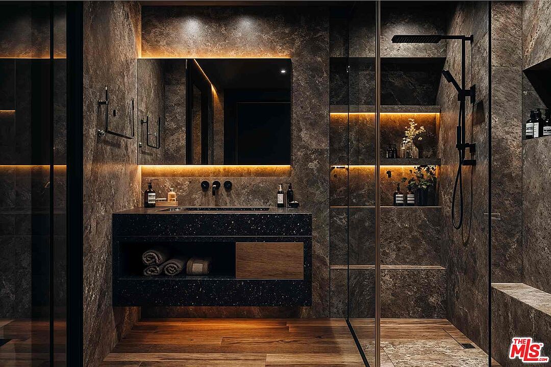 This is a luxurious primary bathroom featuring dark stone walls and wood flooring. The vanity is a dark speckled stone with a built-in sink and storage, illuminated by warm lighting above and below the mirror. A glass-enclosed shower with built-in shelves and a rainfall showerhead adds to the spa-like atmosphere.