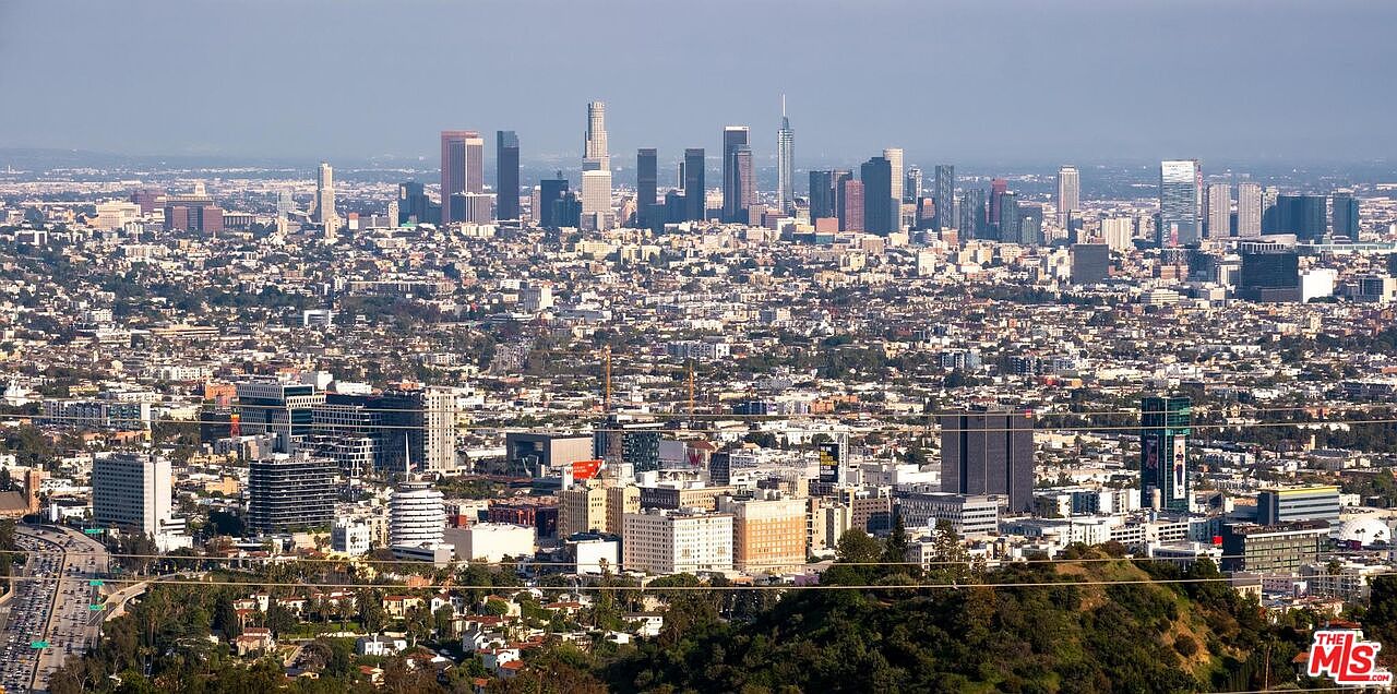 This aerial view showcases the sprawling cityscape of Los Angeles, with a clear focus on the downtown skyline and surrounding urban landscape. The image captures a vast expanse of buildings, streets, and green spaces, offering a comprehensive perspective of the city's density and layout. The composition emphasizes the scale and grandeur of the urban environment, highlighting the mix of residential, commercial, and natural elements.