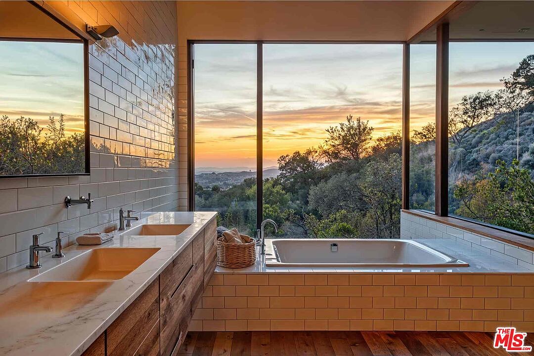 This primary bathroom features a modern design with a double vanity, a soaking tub, and large windows offering stunning views of the surrounding landscape. The bathroom is tiled with white subway tiles and has wood flooring, creating a spa-like atmosphere. The natural light and serene setting enhance the luxurious feel of the space.