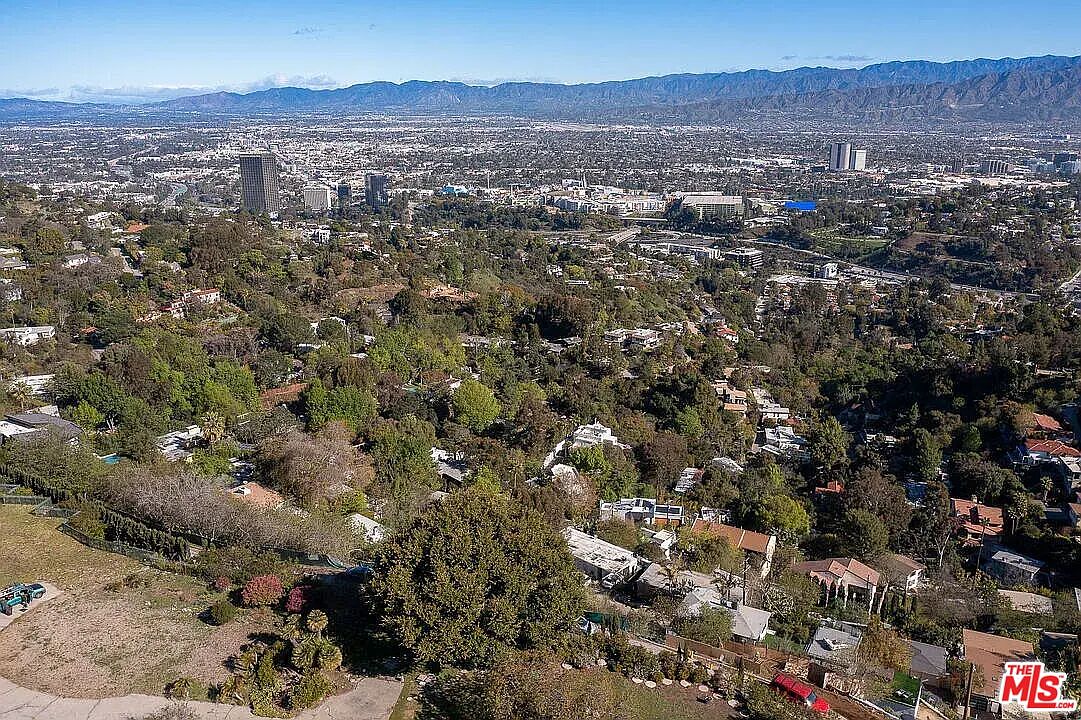 This aerial view showcases a sprawling residential area nestled among lush greenery, with a cityscape and mountains visible in the background. The homes are interspersed with mature trees, creating a sense of privacy and tranquility. The perspective gives a broad overview of the neighborhood's layout and its proximity to both urban amenities and natural landscapes.