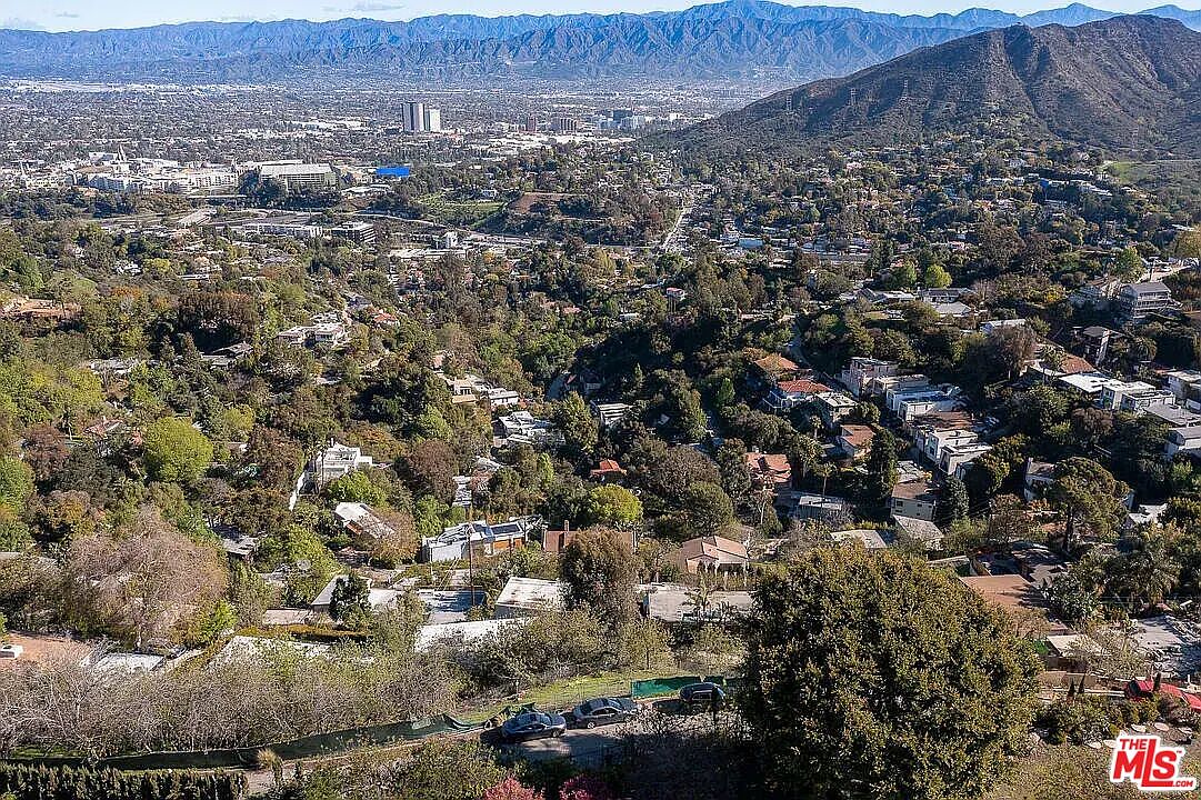 This aerial view showcases a sprawling residential area nestled among lush greenery and rolling hills. The landscape features a mix of houses, trees, and distant mountains, creating a sense of tranquility and seclusion. The perspective provides a comprehensive overview of the neighborhood's layout and natural surroundings.