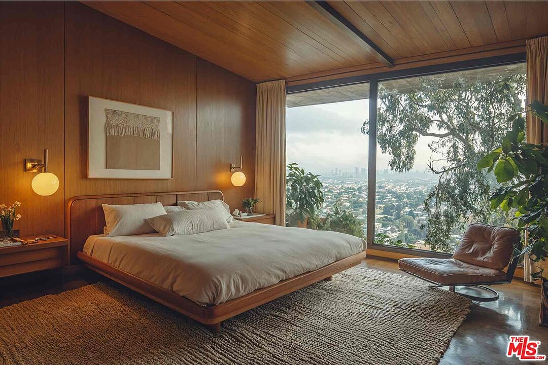 This is a primary bedroom featuring wood paneling and a large window with a city view. The room is furnished with a platform bed, nightstands with globe sconces, and a leather chair. A woven rug adds texture to the space, creating a warm and inviting atmosphere.
