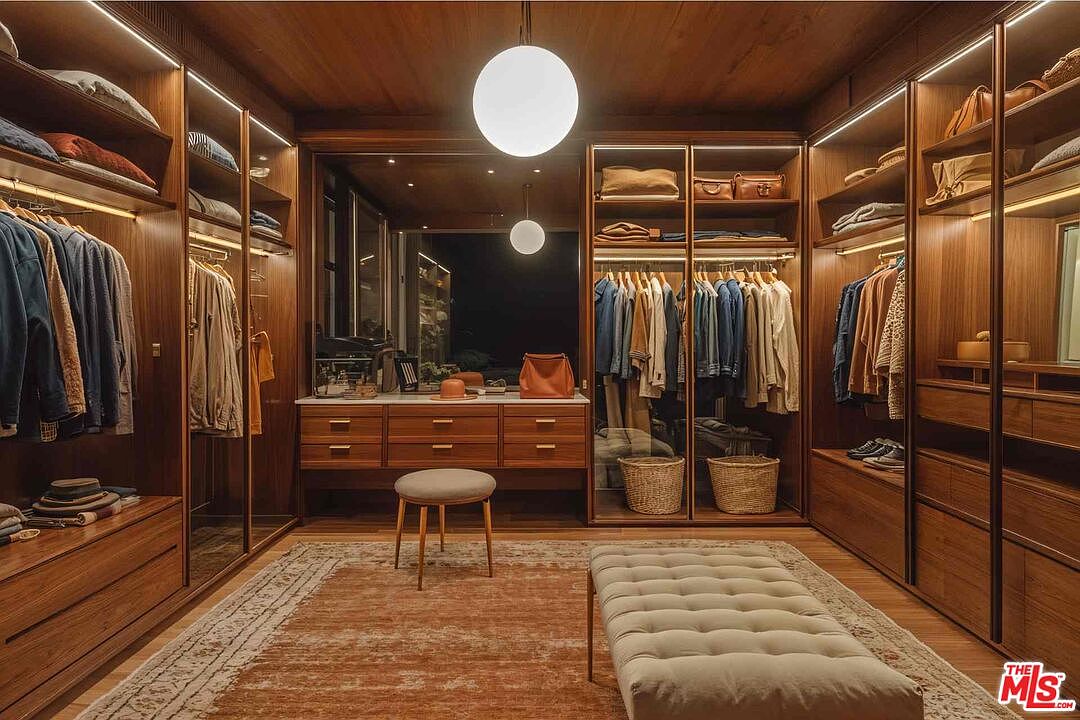 This is a well-organized walk-in closet featuring wooden cabinetry with glass doors and integrated lighting. The closet contains a variety of clothing items, shoes, and accessories, neatly arranged on shelves and hanging rods. A central island with drawers and a seating area adds functionality and style to the space, while a patterned rug covers the floor.