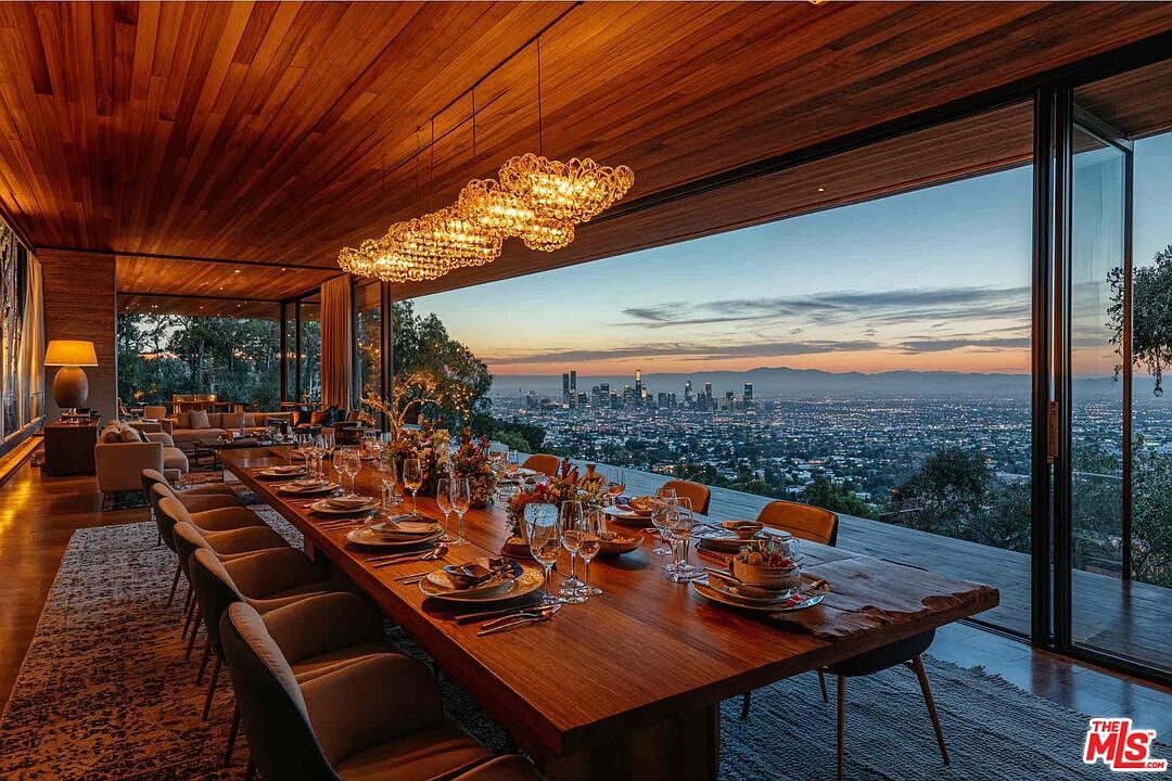 This interior shot showcases a luxurious dining room with a long wooden table set for a formal meal, illuminated by a modern chandelier. Floor-to-ceiling windows offer a breathtaking panoramic view of the city skyline at dusk, creating a sophisticated and elegant ambiance. The room also features a glimpse into a living area, suggesting an open-concept design.