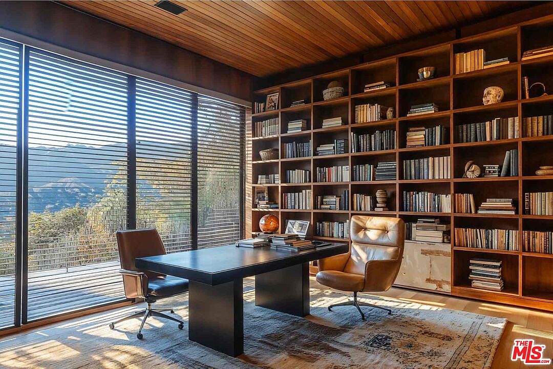 This is an interior shot of a luxurious home office featuring a large wooden bookshelf filled with books, a dark-colored desk with two leather chairs, and a large window with blinds offering a view of the landscape. The room has a warm and sophisticated ambiance, enhanced by the wooden ceiling and the patterned rug, creating an ideal workspace.