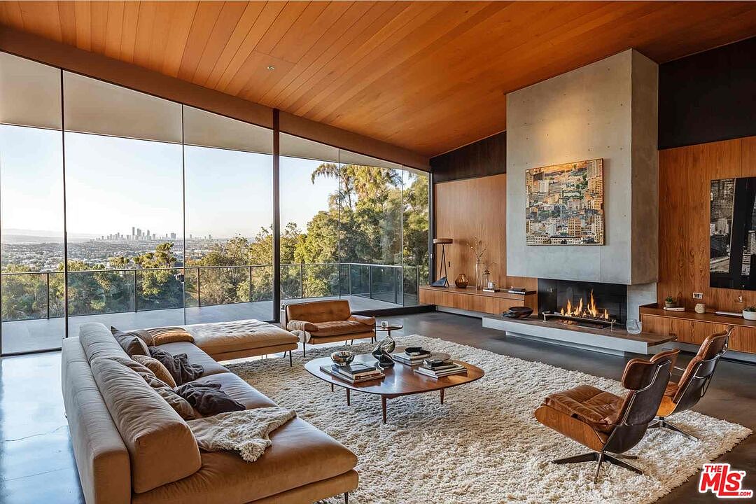 This is a spacious living room featuring a modern design with floor-to-ceiling windows offering a panoramic view of the city and surrounding greenery. The room is furnished with a large sectional sofa, a unique coffee table, and stylish chairs, all set on a plush rug. A striking fireplace with a concrete surround and wood paneling adds a focal point to the room, enhancing the luxurious and comfortable atmosphere.