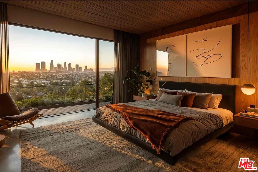 This is a luxurious primary bedroom featuring a platform bed with a dark headboard and a mix of pillows and a throw blanket. The room boasts floor-to-ceiling windows offering a stunning cityscape view, complemented by modern artwork and stylish lighting. The overall impression is one of sophisticated comfort and elegance.