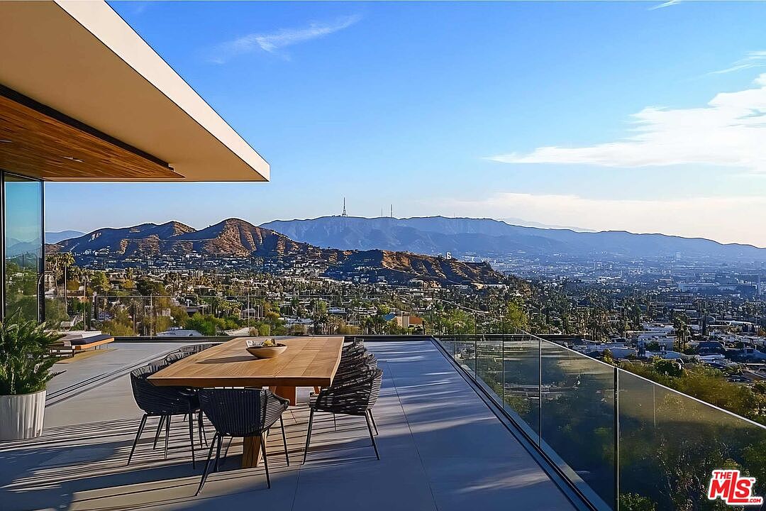 This image showcases a luxurious outdoor patio or deck area with a stunning panoramic view of a city and mountains. A modern dining table with chairs is centrally located, suggesting an ideal space for outdoor dining and entertaining. The patio features a sleek glass railing, enhancing the unobstructed view and adding to the contemporary design of the property.