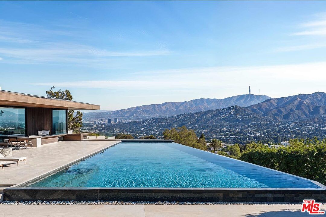This image showcases a luxurious infinity pool with stunning panoramic views of the city and mountains. The pool is surrounded by a modern patio, and a contemporary house with large windows is visible on the left. The clear blue sky and serene water create an inviting and upscale atmosphere.