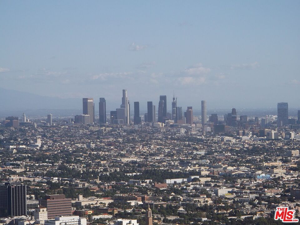 This aerial view showcases a sprawling cityscape under a clear, blue sky. Prominent skyscrapers punctuate the horizon, set against a backdrop of densely populated urban areas. The image conveys a sense of vastness and urban density, highlighting the scale of the city.