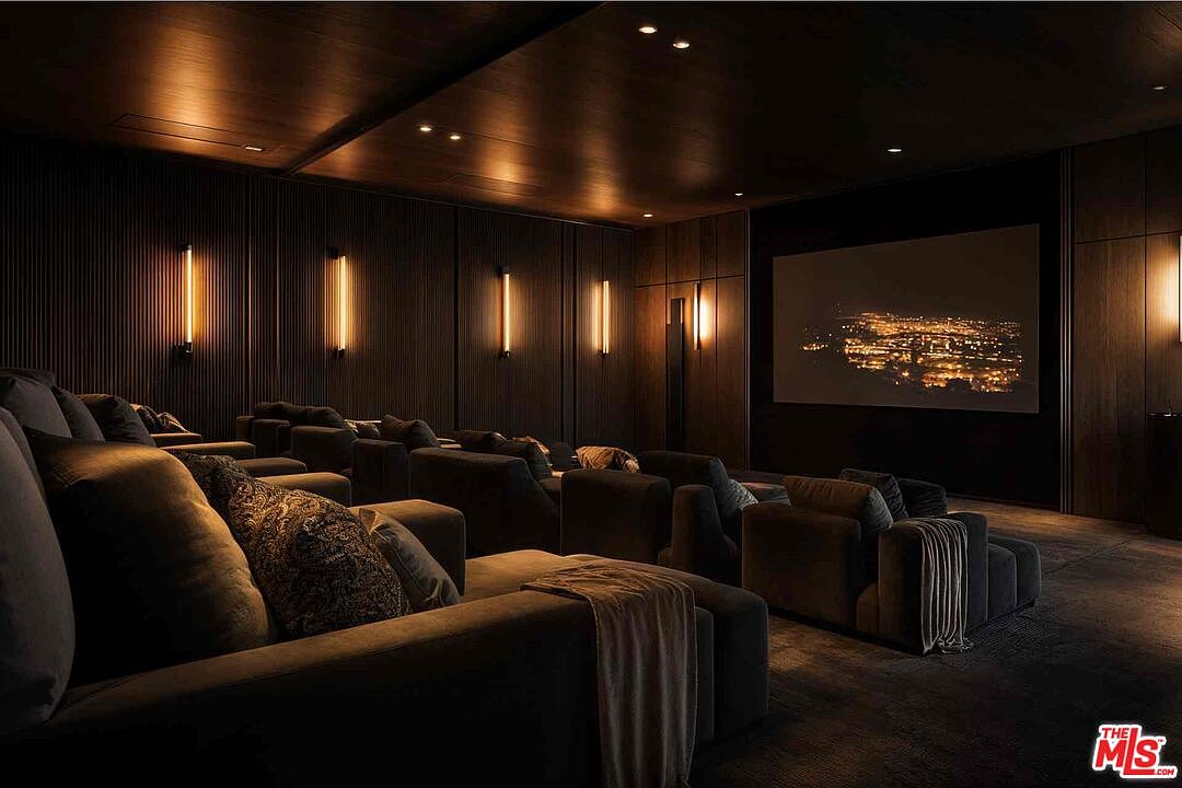This is a luxurious home theater featuring tiered seating with plush, dark-colored sofas and decorative pillows. The walls are adorned with vertical light fixtures, creating a warm and inviting ambiance. A large screen displays a cityscape at night, enhancing the cinematic experience.