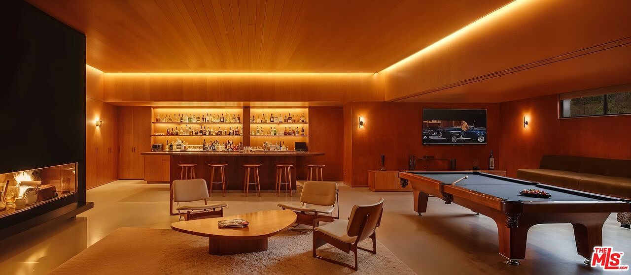 This is a warm and inviting basement space featuring a bar with liquor shelves, a pool table, and a seating area with a unique coffee table. The room is illuminated by recessed lighting and wall sconces, creating a cozy atmosphere. The wood paneling and neutral color palette contribute to a sophisticated and relaxing environment.