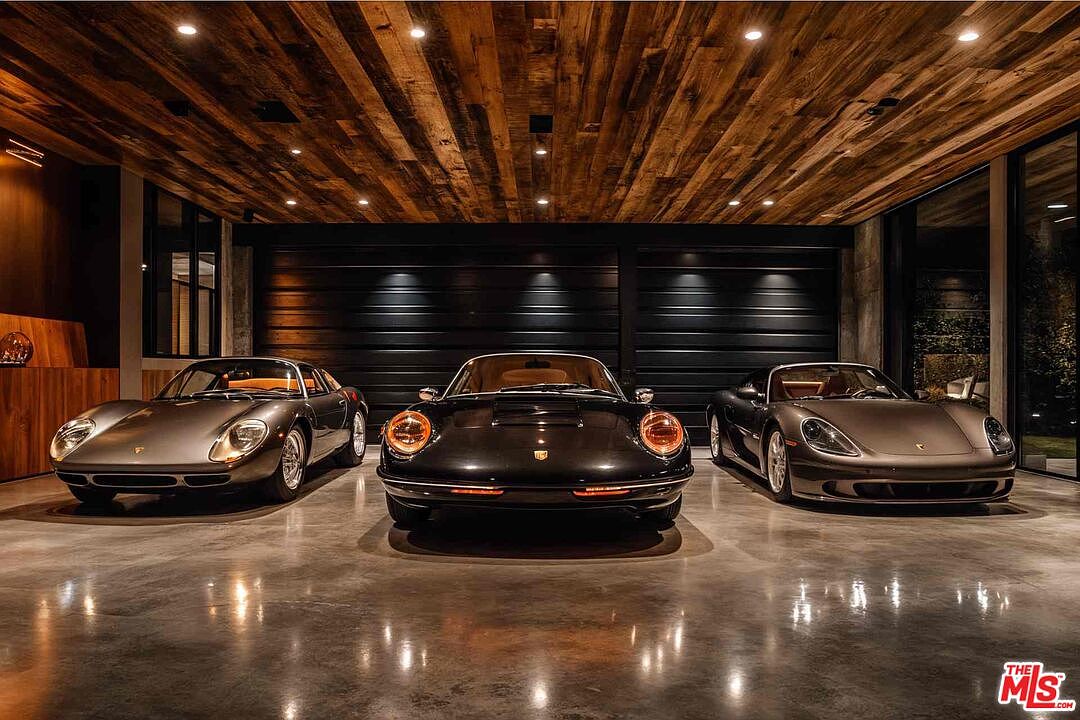 This image showcases a luxurious garage featuring three classic cars parked on a polished concrete floor. The garage has a modern design with a wooden plank ceiling and sleek, dark garage doors. Large windows on either side of the garage doors provide natural light and a view of the exterior.