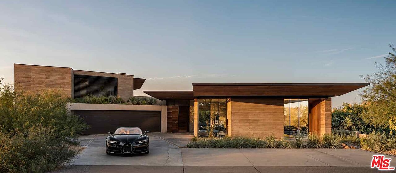 This is a front exterior view of a modern, luxurious home. The architecture features clean lines, wood and concrete elements, and large glass windows. A black Bugatti Chiron is parked in the driveway, adding to the property's high-end appeal. The landscaping is minimal and complements the contemporary design of the house.