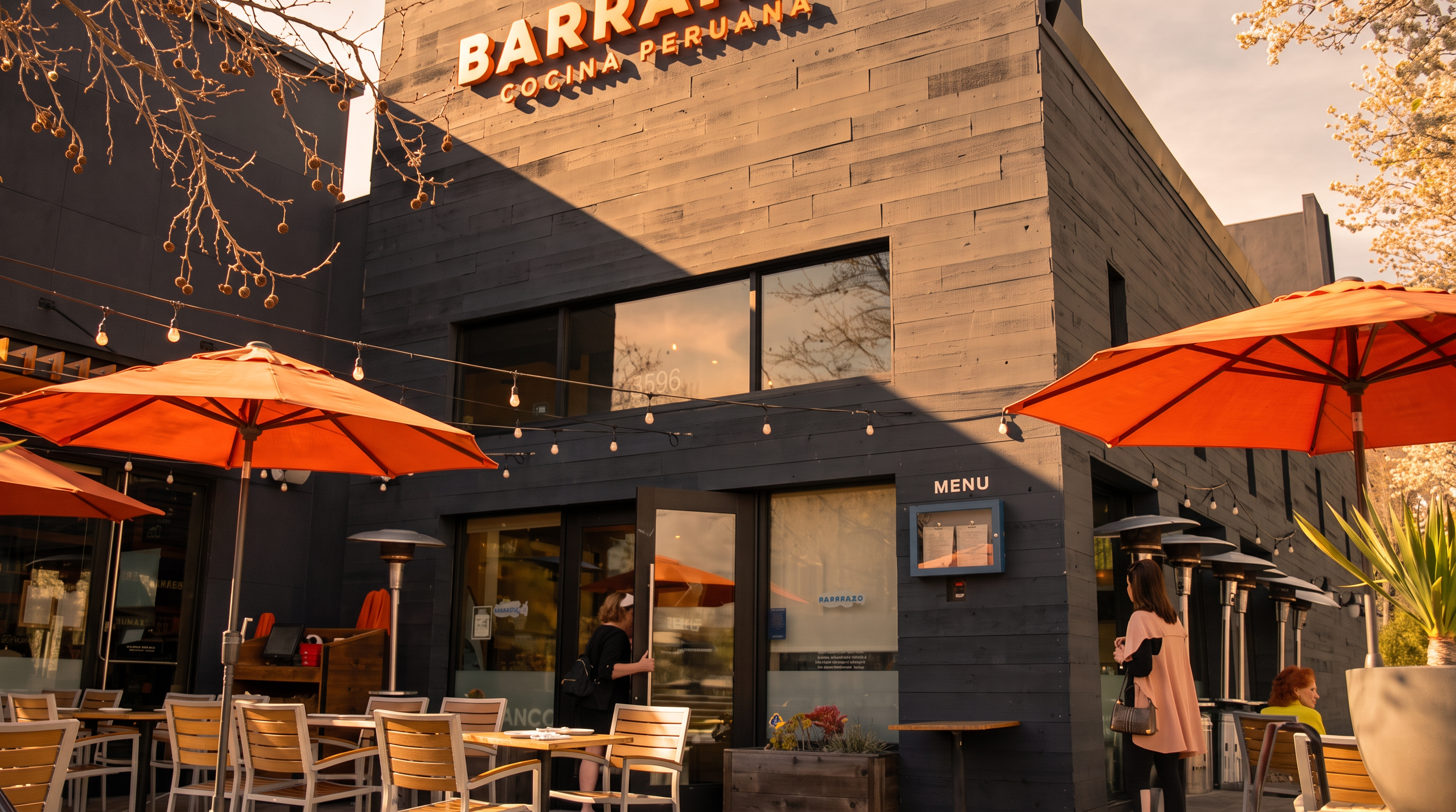 This image shows the exterior facade of Barranco, a Peruvian restaurant featuring a dark, modern horizontal wood-plank siding and large windows. The establishment provides an inviting outdoor dining space with multiple patio tables, chairs, and orange umbrellas. The scene is enhanced by warm string lighting, giving the restaurant a welcoming and vibrant urban atmosphere.