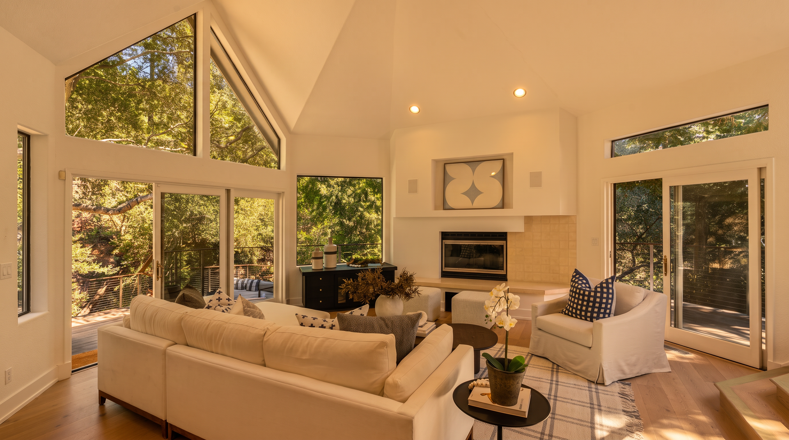 This bright and airy living room features a vaulted ceiling and large windows that provide expansive views of the surrounding trees. The space is anchored by a fireplace with a light-colored tile surround and a comfortable seating area consisting of a white sectional sofa and a plush armchair. Light wood flooring and a neutral color palette create a serene, modern atmosphere that seamlessly connects the indoor living space with the outdoor deck.