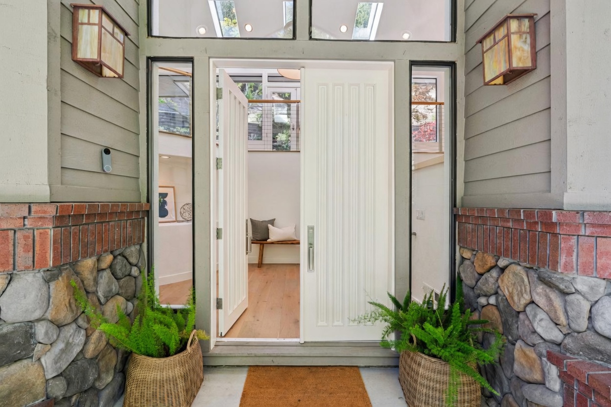 This inviting entryway features a modern white front door with vertical paneling, flanked by tall, narrow windows that allow natural light to filter into the home. The exterior is framed by stone-clad walls and horizontal siding, accented by two warm-toned wall sconces and lush potted ferns on either side of the entrance. A welcome mat sits on the porch, providing a warm and professional first impression of the property.
