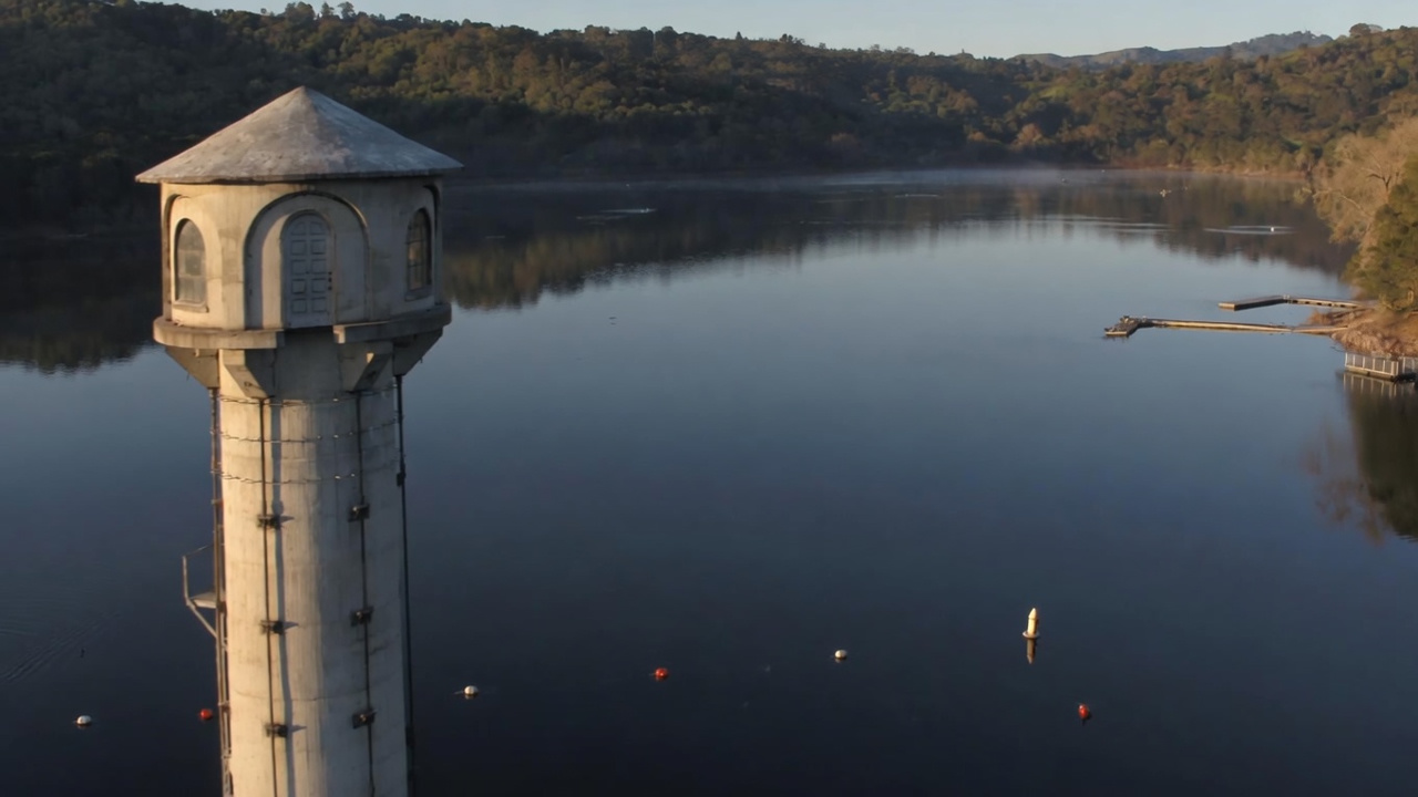 This scenic image captures a prominent, cylindrical stone water tower rising from a calm, reflective reservoir. The tranquil water is marked by a series of buoys and a floating dock extending from the shoreline, suggesting a peaceful recreational area for the community. The surrounding hilly, wooded landscape enhances the natural and serene aesthetic of this unique landmark.