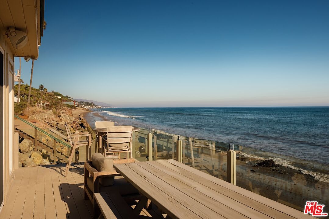 This image showcases a beautiful outdoor deck overlooking the ocean. The deck features wooden flooring, a glass railing, and outdoor furniture including a picnic table and chairs. The view extends to the beach and the ocean, creating a serene and inviting atmosphere.