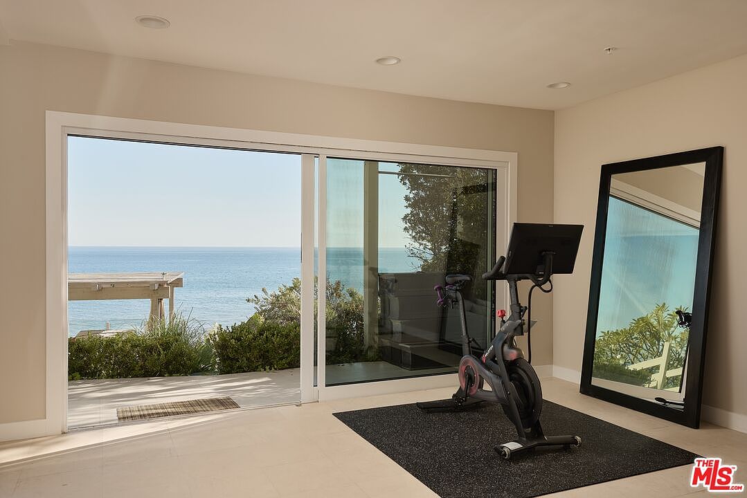 The image showcases a well-lit home gym with a stunning ocean view through a sliding glass door. A stationary bike sits on a black mat, positioned near a full-length mirror, creating a dedicated workout space. The room features neutral-toned walls and tile flooring, contributing to a clean and inviting atmosphere.