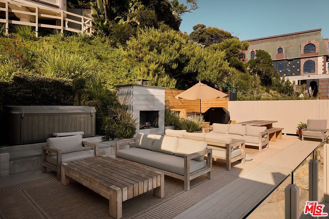 This is a well-appointed outdoor patio area, featuring comfortable seating arrangements with sofas and chairs, a wooden coffee table, and a dining table with benches. A modern fireplace and a hot tub add to the luxurious feel, while an umbrella provides shade. The space is surrounded by lush greenery and a wooden fence, creating a private and inviting atmosphere.