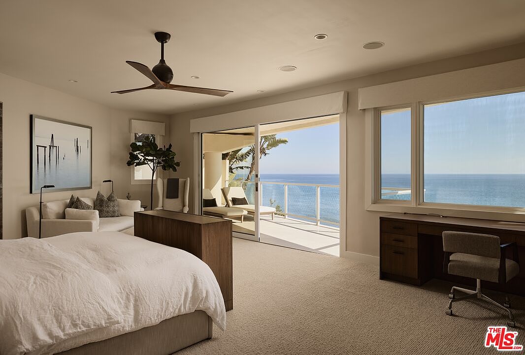 This is a bright and airy primary bedroom with a view of the ocean. The room features a large bed with white linens, a seating area with a sofa, and a desk area with a chair. Sliding glass doors lead to a balcony, and a ceiling fan provides air circulation. The overall style is modern and luxurious.