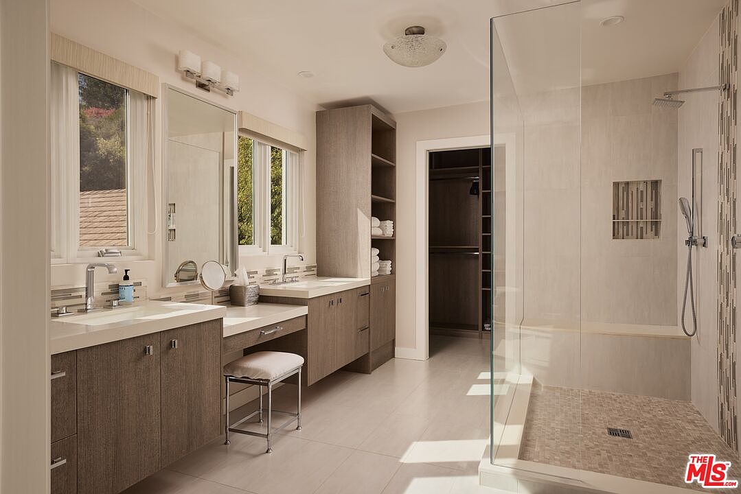 This is a well-lit primary bathroom featuring a double vanity with modern fixtures and a makeup area with a stool. A glass-enclosed shower with a built-in bench and tiled walls adds a luxurious touch. The color palette is neutral, creating a serene and spa-like atmosphere.