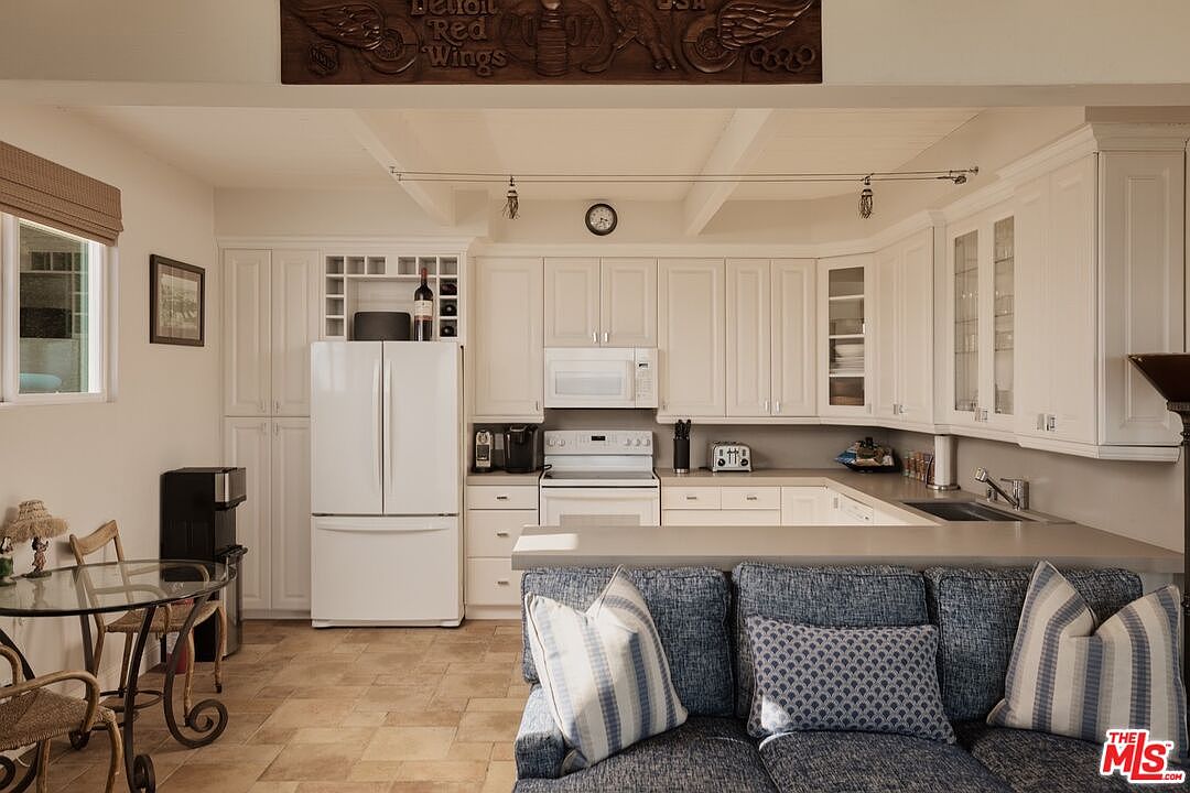 This is a well-lit kitchen featuring white cabinetry, stainless steel appliances, and a neutral-toned countertop. The kitchen includes a refrigerator, microwave, oven, and a wine rack built into the cabinetry. A partial view of a blue sofa with decorative pillows suggests an open-concept living space adjacent to the kitchen.