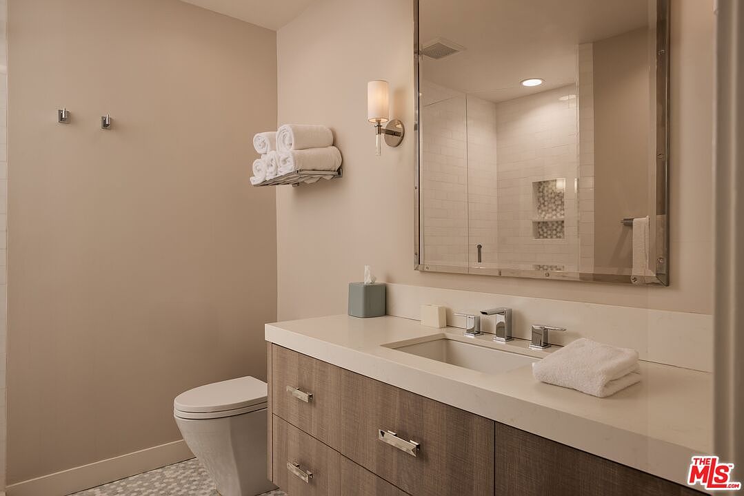 This is a well-lit bathroom featuring a modern vanity with a light wood-grain finish and a white countertop. A large mirror reflects the shower area, and the neutral color palette creates a clean and serene atmosphere. The toilet is a modern, low-profile design.