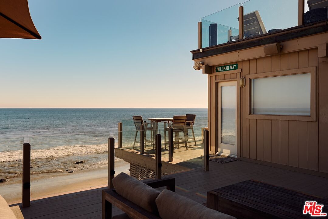 This image showcases a stunning oceanfront patio/deck area, perfect for relaxation and entertainment. The deck features comfortable seating, a dining table with chairs, and a glass railing offering unobstructed views of the ocean. The property's exterior is visible, with a sign reading 'Wildman Way,' adding a touch of character to this coastal retreat.