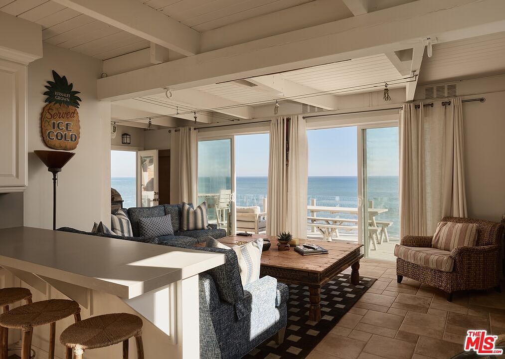 This is an interior shot of a living room with a coastal vibe, featuring a partial view of the kitchen counter and stools in the foreground. The living area includes a comfortable blue patterned sofa and armchair, a rustic wooden coffee table, and large sliding glass doors that offer stunning ocean views. The room is well-lit with natural light, enhancing the relaxed and airy atmosphere.