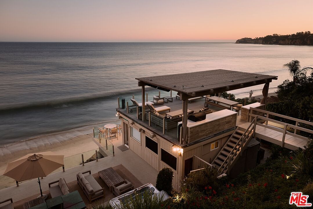 This image showcases a multi-level deck and patio area of a beachfront property, offering stunning ocean views. The upper deck features a covered lounge area with comfortable seating, while the lower level includes a dining set and additional lounge furniture, creating an inviting outdoor living space. A staircase and walkway connect the deck to the surrounding landscape, blending seamlessly with the natural environment.