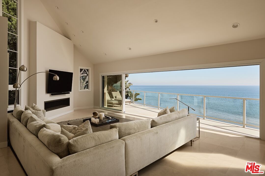 This is a bright and airy living room with a stunning ocean view. The room features a large sectional sofa, a modern fireplace with a mounted television, and floor-to-ceiling windows that open onto a balcony. The neutral color palette and minimalist decor create a serene and luxurious atmosphere.